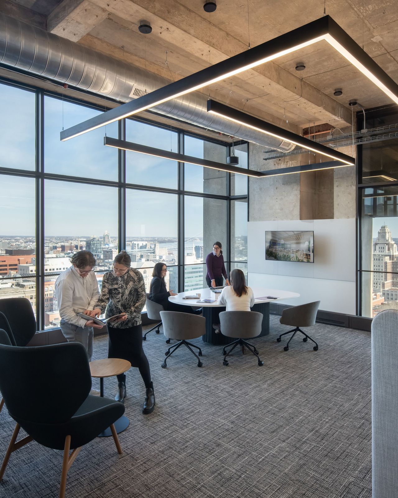 Modern office interior with floor-to-ceiling windows overlooking a city skyline, featuring a small meeting area with people gathered around a table, contemporary lounge seating, exposed ceilings, and linear pendant lighting.